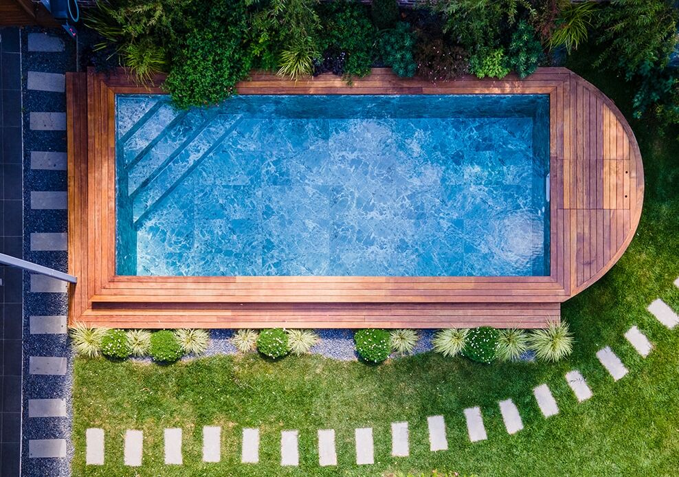 Vue aérienne d'une piscine rectangulaire dotée d'une terrasse en bois, entourée d'une pelouse verdoyante, de pierres de gué et de diverses plantes le long des bords : une oasis accueillante qui met en valeur l'élégance caractéristique des piscines modernes.