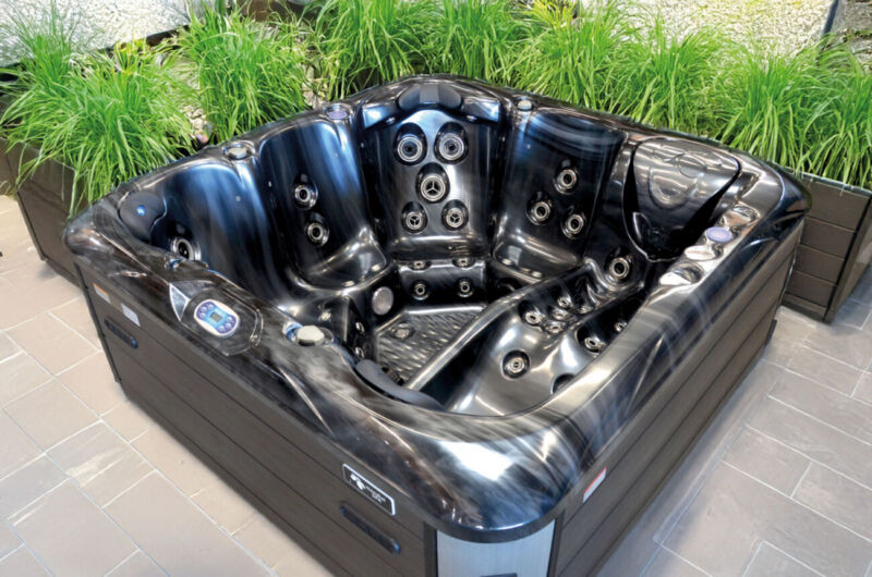 Un jacuzzi moderne, noir et gris, doté de multiples jets et de commandes numériques, trône sur un sol carrelé, entouré de plantes vertes disposées dans des jardinières rectangulaires, offrant un havre de paix luxueux qui rappelle les piscines haut de gamme.