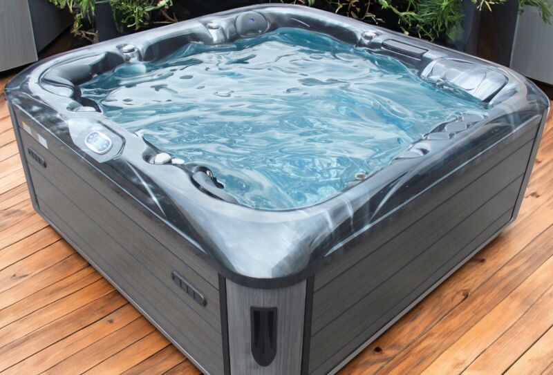 Un jacuzzi moderne, de couleur grise, rempli d'eau limpide, trône sur une terrasse en bois, entouré de plantes vertes en arrière-plan, créant ainsi une atmosphère accueillante qui rappelle celle des piscines de luxe.