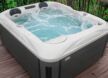 Un jacuzzi bouillonnant aux bords blancs est installé sur une terrasse en bois, entouré de verdure et d'une balustrade noire.