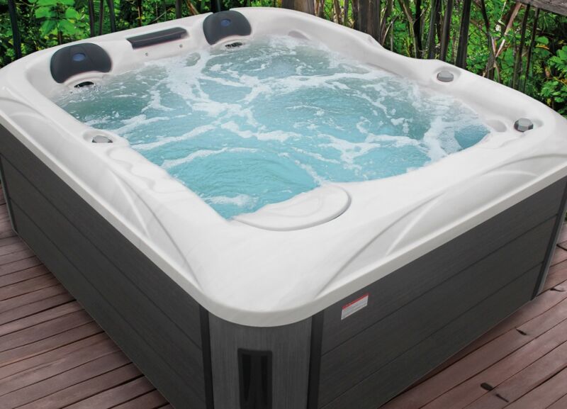 Un jacuzzi bouillonnant aux bords blancs est installé sur une terrasse en bois, entouré de verdure et d'une balustrade noire.