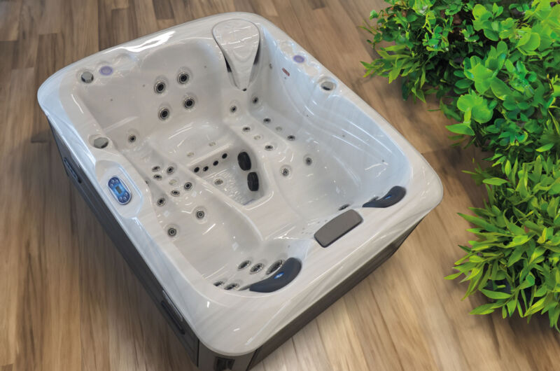 Un jacuzzi blanc pour deux personnes, doté de plusieurs jets et qui rappelle les piscines de luxe, trône sur un parquet à côté d'une plante aux feuilles vertes.